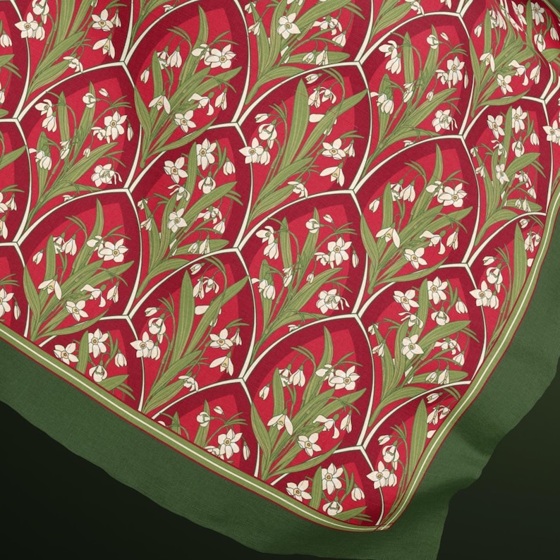 May include: A square fabric scarf featuring a repeating pattern of white snowdrop flowers with green leaves set against a deep red background. The design is organised into arched, scale-like segments outlined in white, finished with a solid green border.