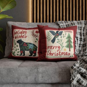 May include: Two square throw pillows with deep red borders and cream-colored centers. One pillow features a black bear with floral patterns and the text "holiday wishes". The other displays a bird, a green pine tree, and "merry Christmas".