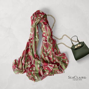May include: A long scarf featuring a deep red and olive green floral pattern with white blossoms, paired with a small dark green handbag that has a gold-toned metal chain strap and a gold-toned clasp. The text SEACLAIRE copyright 2026 is visible.