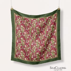 May include: A square scarf featuring a repeating pattern of white flowers and green leaves set within deep red arched segments. The scarf has a solid dark green border and is suspended by two metal clips. The text Seaclaire copyright 2026 is visible.