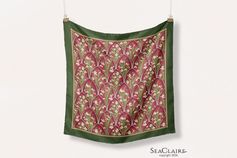 May include: A square scarf featuring a repeating pattern of white flowers and green leaves set within deep red arched segments. The scarf has a solid dark green border and is suspended by two metal clips. The text Seaclaire copyright 2026 is visible.