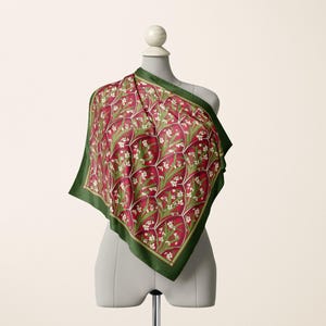 May include: A square scarf featuring a deep red background with a repeating pattern of white flowers and green leaves, enclosed by a dark green border. The scarf is draped over a mannequin, displaying a geometric grid design across the fabric surface.