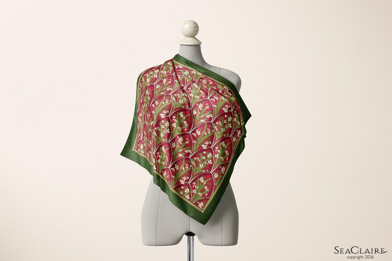 May include: A square scarf featuring a deep red background with a repeating pattern of white flowers and green leaves, enclosed by a dark green border. The scarf is draped over a mannequin, displaying a geometric grid design across the fabric surface.