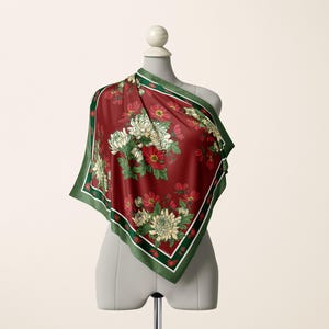 May include: A square scarf featuring a deep red background with a floral pattern of white and red chrysanthemums and green leaves. The scarf has a dark green border with thin white and red stripes along the edges, draped over a mannequin torso.