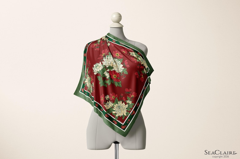 May include: A square scarf featuring a deep red background with a floral pattern of white and red chrysanthemums and green leaves. The scarf has a dark green border with thin white and red stripes along the edges, draped over a mannequin torso.