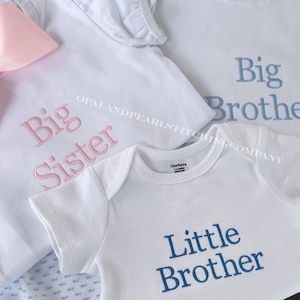 May include: Three white garments including a shirt with pink embroidered text reading Big Sister, a shirt with blue embroidered text reading Big Brother, and a onesie with blue embroidered text reading Little Brother, plus a pink bow accessory.