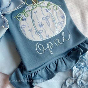 May include: A dusty blue shirt featuring an embroidered pumpkin with a white and blue floral ribbon pattern. The pumpkin has a brown stem and green vines. The name Opal is embroidered in white cursive below the pumpkin. The shirt has a ruffled hem.