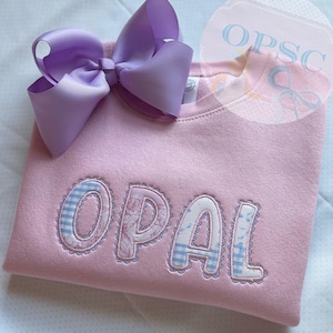 May include: A light pink sweatshirt featuring the name OPAL appliqued in a mix of blue and white striped fabric and a floral pattern. The letters are outlined with decorative stitching. A large lavender ribbon bow is attached to the neckline.