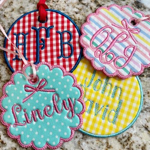 May include: Four embroidered fabric tags with scalloped edges and ribbons for hanging. Designs include a red and white gingham tag with blue initials, a striped tag with pink initials, a teal polka dot tag with the name "Linely", and a yellow gingham tag with the names "John David".