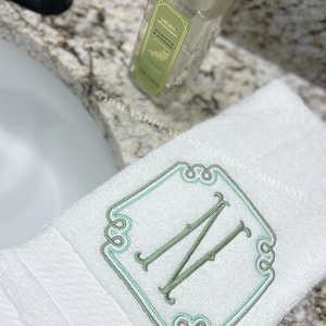 May include: A white hand towel featuring an embroidered sage green letter N centered within a decorative sage green and light teal frame. The towel has a textured border and the text OPALANDPEARLSTITCHINGCOMPANY is visible across the fabric.