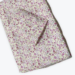 May include: A floral bedding set featuring a fitted sheet and pillowcase. The pattern displays small purple and pink flowers with green leaves on a cream backdrop. The fabric is folded, suggesting a soft texture. The set is designed for a bed.