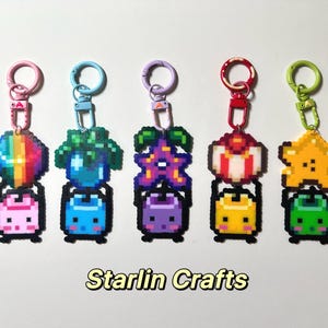 May include: Five pixel art keychains with colourful designs. Each keychain features a charm with a unique shape and colour, such as a rainbow sphere, a blue plant, a purple flower, a red and white dessert, and a yellow star. The keychains have a metal clasp and the text "Starlin Crafts".
