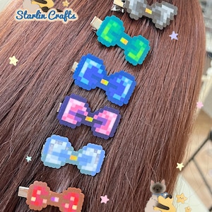 May include: Six pixelated bow hair clips in various colors, including blue, green, pink, and brown, are displayed against a backdrop of brown hair. Each clip has a metal clasp. The Starling Crafts logo is visible in the image.