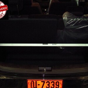 May include: A white automotive rear strut tower bar installed horizontally across the cargo area of a vehicle. The metal bar features mounting brackets at each end and a small rectangular label in the center. A red circular graphic in the corner displays the text IMPORT TAX INCLUDED IMPORT.