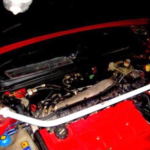 May include: A white strut bar installed in a car engine bay. The engine bay is red and black, with various mechanical components visible. The strut bar spans across the engine bay, connecting the strut towers for enhanced stability.