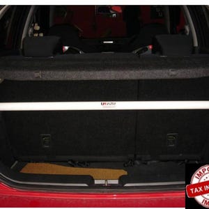 May include: A white horizontal metal strut bar installed across the interior cargo area of a red vehicle. The bar features a small logo with the text Ultra Racing printed in the center, positioned against the black fabric of the rear seat back.
