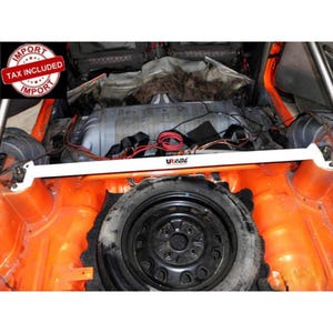 May include: A white metal rear strut bar with the Ultra Racing logo installed in the trunk of a vehicle with a bright orange interior. The bar is positioned horizontally above a black spare tire, with visible wiring and metal components.
