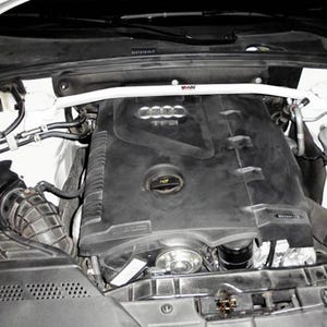 May include: An overhead view of a car engine bay. The engine is black with a white strut bar across the top. The engine bay is white with black components and hoses. The engine has the Audi logo and the letters T F S I.
