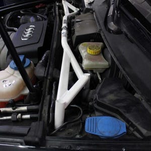 May include: A white strut bar installed in a car engine compartment. The engine bay is black, with various components visible, including an Audi logo. A person is working on the car with a wrench.