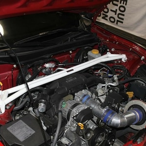 May include: A detailed view of a car engine bay. The engine is red with a white strut bar and various black and silver components. Blue tubing and a battery are also visible.
