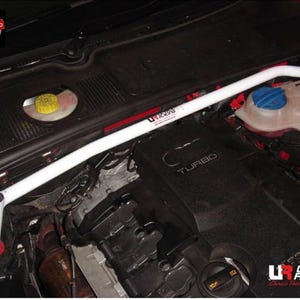May include: A white metal strut bar installed across the engine bay of a vehicle. The bar features the Ultra Racing logo in black and red text. The engine compartment includes a black engine cover with the word Turbo and a yellow fluid cap.