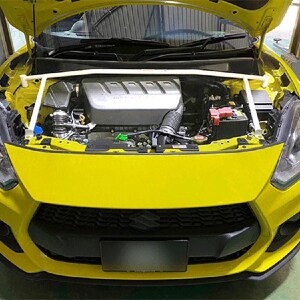 May include: Bright yellow car with the hood open, revealing the engine compartment. A white strut bar spans the engine bay. The car's front features a black grille and headlights. The engine is silver and black.