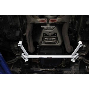 May include: A white automotive undercarriage brace bar installed beneath a vehicle chassis. The metal component features a central horizontal bar with angled support arms on each side, secured with bolts, and includes small branding labels on the center.