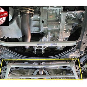 May include: A light gray metal automotive undercarriage brace bar installed beneath the engine and transmission components of a vehicle. The structural reinforcement bar features a tubular design with visible mounting points and a metallic finish.