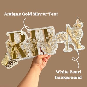 May include: A decorative sign spelling "RITA" in antique gold mirror text, set against a white pearl background. The design incorporates gold floral and butterfly accents, offering an elegant and personalized aesthetic.