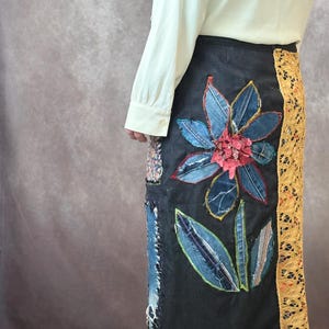 May include: Black denim skirt with a large, appliquéd flower design. The flower features blue denim petals and a red center. A yellow lace panel runs down the side. Patches of fabric are sewn on the skirt.