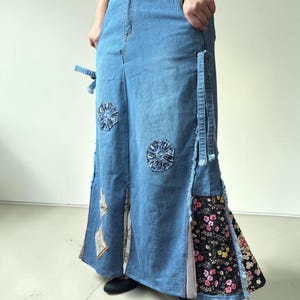 Puede incluir: Una falda larga de mezclilla azul con cintura alta, bolsillos laterales y bordes deshilachados. La falda está adornada con parches circulares de patrones azules y un panel lateral grande de tela negra con estampado floral.