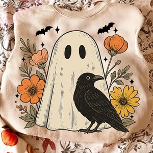 May include: A cream-colored sweatshirt featuring a vintage-style Halloween design. The graphic includes a friendly ghost, a black crow, orange and yellow flowers, and bats. The design is surrounded by floral elements and stars.