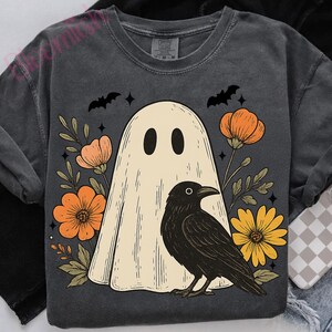 May include: Dark gray sweatshirt featuring a cream-colored ghost with black eyes, a black crow, and orange and yellow flowers. Two black bats and stars are above the ghost. The design has a vintage, slightly faded look.