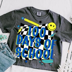May include: Dark gray t-shirt with the text "100 Days of School" in blue and yellow, set against a black and white checkered background. The design includes a yellow pencil, a smiley face, stars, lightning bolts, a skateboard, and a skeleton hand.