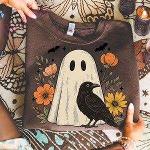 May include: Brown sweatshirt featuring a cream-colored ghost with black eyes, a black crow, and orange and yellow flowers. Two black bats and stars are above the ghost. The sweatshirt is held by two hands.