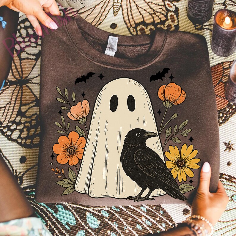 May include: Brown sweatshirt featuring a cream-colored ghost with black eyes, a black crow, and orange and yellow flowers. Two black bats and stars are above the ghost. The sweatshirt is held by two hands.