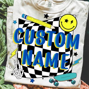 May include: Cream-colored t-shirt with a graphic design featuring a checkered pattern, a yellow pencil, a smiley face, and the words "CUSTOM NAME" in blue and yellow. Additional graphics include stars, lightning bolts, a peace sign, and a skateboard.