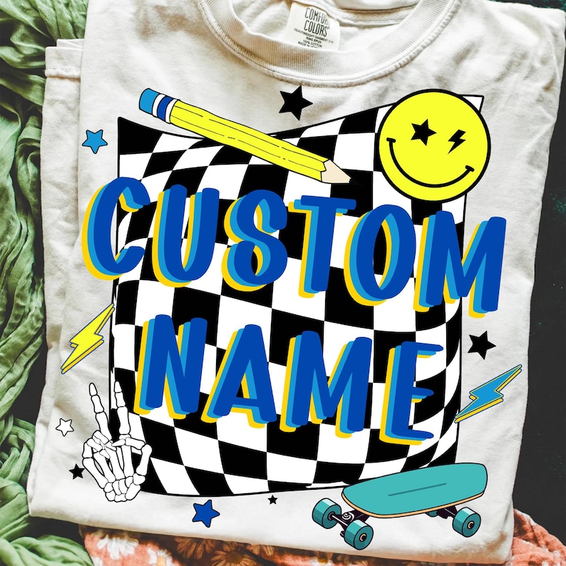 May include: Cream-colored t-shirt with a graphic design featuring a checkered pattern, a yellow pencil, a smiley face, and the words "CUSTOM NAME" in blue and yellow. Additional graphics include stars, lightning bolts, a peace sign, and a skateboard.