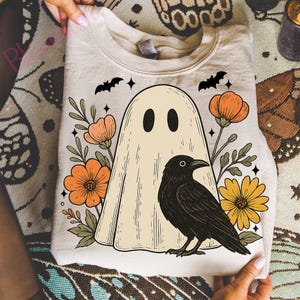 May include: A beige sweatshirt featuring a ghost design with black eyes, surrounded by orange and yellow flowers, a black crow, and bats. The design has a vintage, Halloween theme.
