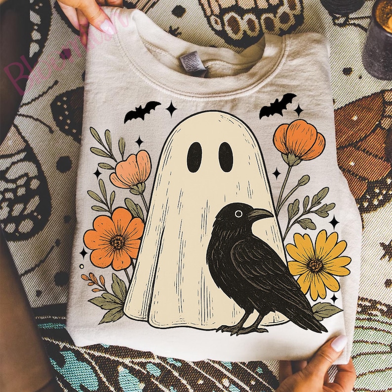 May include: A beige sweatshirt featuring a ghost design with black eyes, surrounded by orange and yellow flowers, a black crow, and bats. The design has a vintage, Halloween theme.