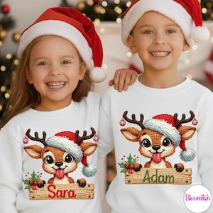 Peut inclure: Deux enfants portant des sweat-shirts blancs avec un motif de renne et des bonnets de Père Noël. Le renne a un bonnet de Père Noël rouge, des bois bruns et tire la langue. Un sweat-shirt dit "Sara" et l'autre "Adam" sur un panneau en bois.