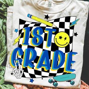 May include: A light-colored t-shirt with a graphic design for 1st grade. The design includes a checkered flag, a yellow pencil, a smiley face, lightning bolts, stars, a skeleton hand, and a skateboard. The text "1st Grade" is in blue with a yellow outline.