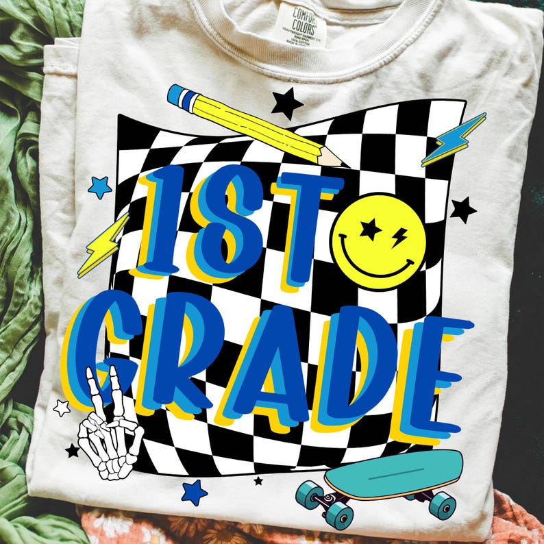 May include: A light-colored t-shirt with a graphic design for 1st grade. The design includes a checkered flag, a yellow pencil, a smiley face, lightning bolts, stars, a skeleton hand, and a skateboard. The text "1st Grade" is in blue with a yellow outline.