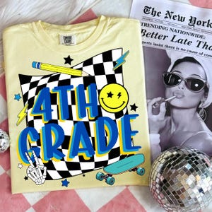 May include: A light yellow Comfort Colors t-shirt with a graphic design for 4th grade. The design includes a checkered flag, a smiley face, a pencil, stars, a skateboard, and the words "4TH GRADE" in blue and yellow.