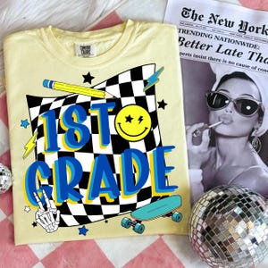 May include: A light yellow t-shirt with a graphic design for 1st grade. The design includes a checkered flag, a smiley face, a pencil, a skateboard, and the words "1st Grade" in blue and yellow. A newspaper is in the background.