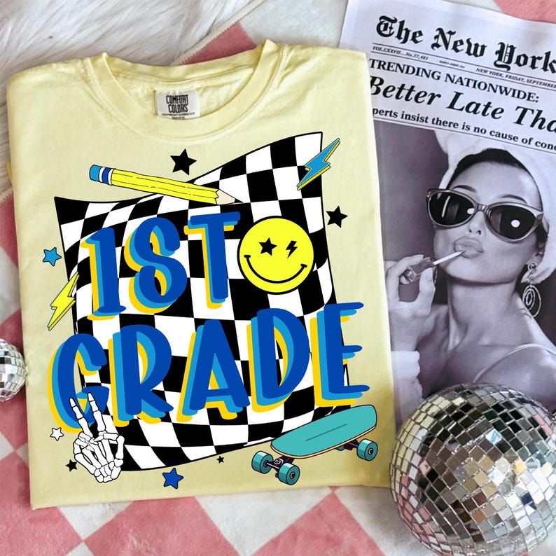May include: A light yellow t-shirt with a graphic design for 1st grade. The design includes a checkered flag, a smiley face, a pencil, a skateboard, and the words "1st Grade" in blue and yellow. A newspaper is in the background.