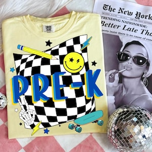 May include: Yellow Comfort Colors t-shirt with a graphic design featuring a checkered flag, a smiley face, a pencil, stars, lightning bolts, a skateboard, and the word "PRE-K" in blue and yellow.