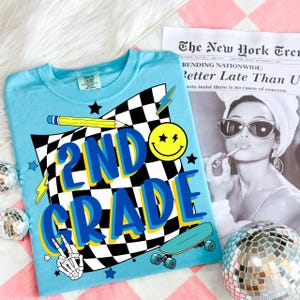 May include: A light blue t-shirt with a graphic design for 2nd grade. The design includes a checkered flag, a yellow smiley face, a pencil, a skateboard, and the words "2ND GRADE" in blue and yellow. The shirt is on a pink and white checkered surface.