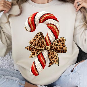 May include: A cream-colored sweatshirt featuring a large, whimsical candy cane design. The candy cane is white, red, and gold, with a leopard print bow. The design is festive and eye-catching, perfect for the holiday season.