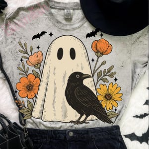 May include: A gray t-shirt featuring a ghost design with a black raven, orange flowers, and yellow flowers. The ghost has two black oval eyes. The shirt also has black bats and stars.